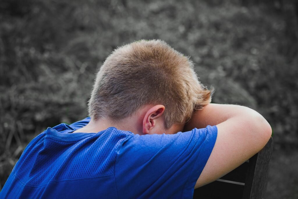 A young boy in a blue shirt rests his head on his arms outdoors, appearing sad and contemplative.